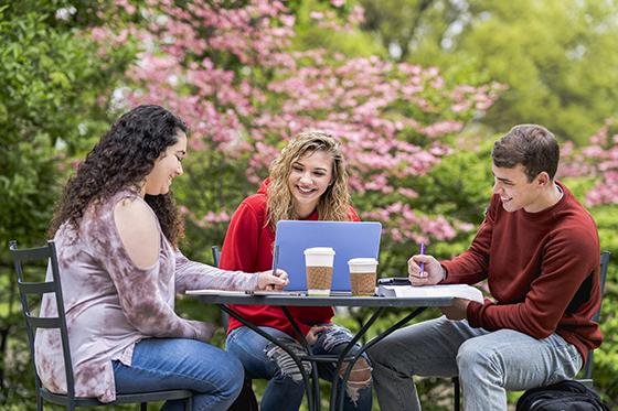 Photo of three Chatham University students sitting at a patio table outside working together on laptops and smiling. 
