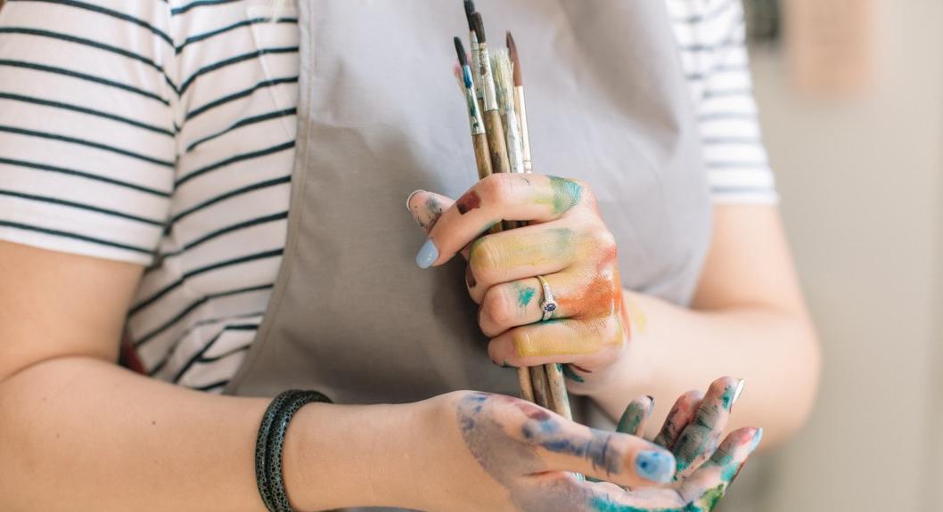 Close-up photo of a Chatham University art student wearing an apron and holding paintbrushes in hands that are covered in paint. 